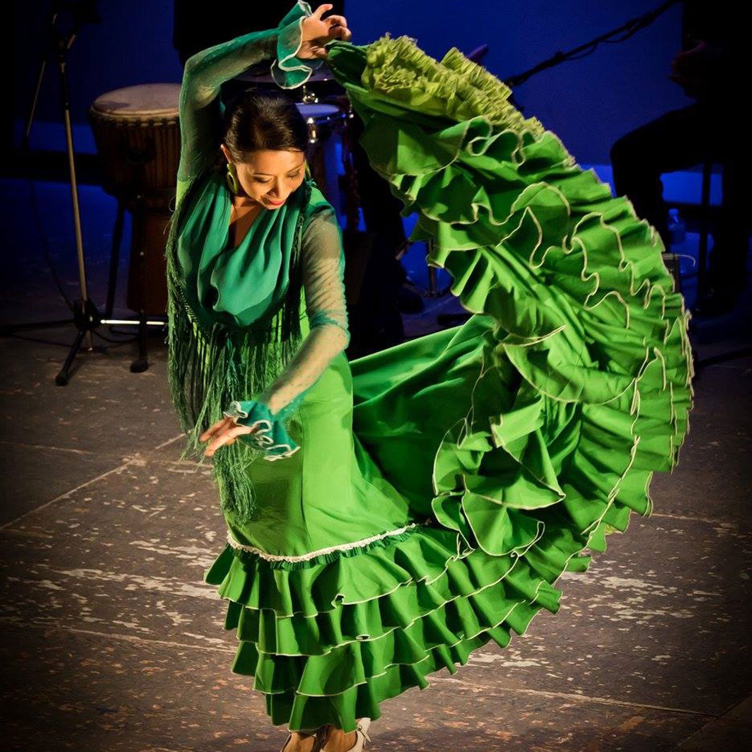 A flamenco dance artist posest on a stage. They are wearing a green traditional Flamenco dress with a long ruffled skirt. They are holding the skirt up above their head with their right hand and are looking down at the left arm which is slightly extended.