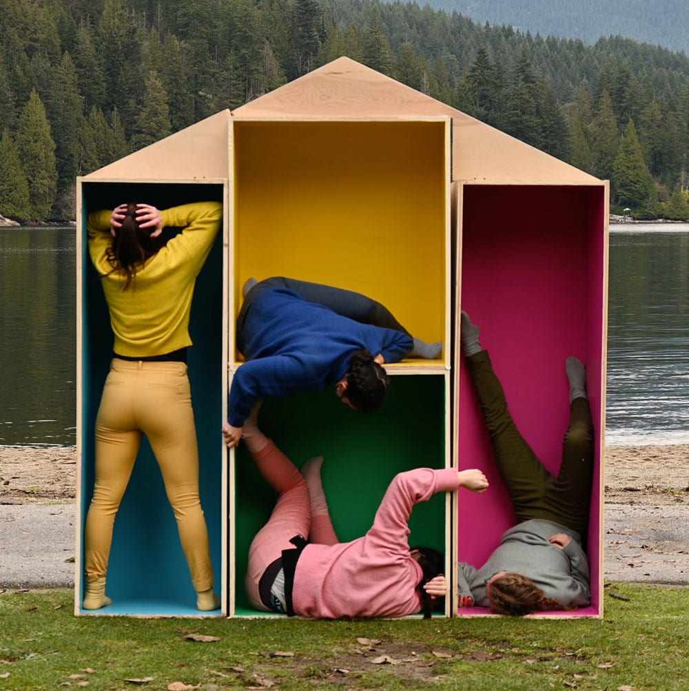 Dance artists pose inside makeshift shelves shaped in a house on a river bank