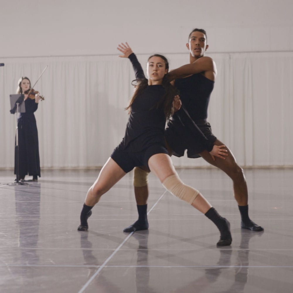 Dance artist Xin Hui Ong posing mid rehearsal with partner while violinist plays behind them