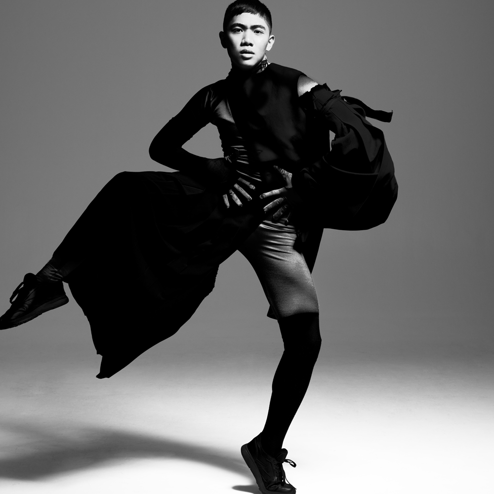 A dance artist poses with one leg extended outward. They have their hands on their waist and are wearing all black with flowing fabric draped down one leg. This image is in greyscale.