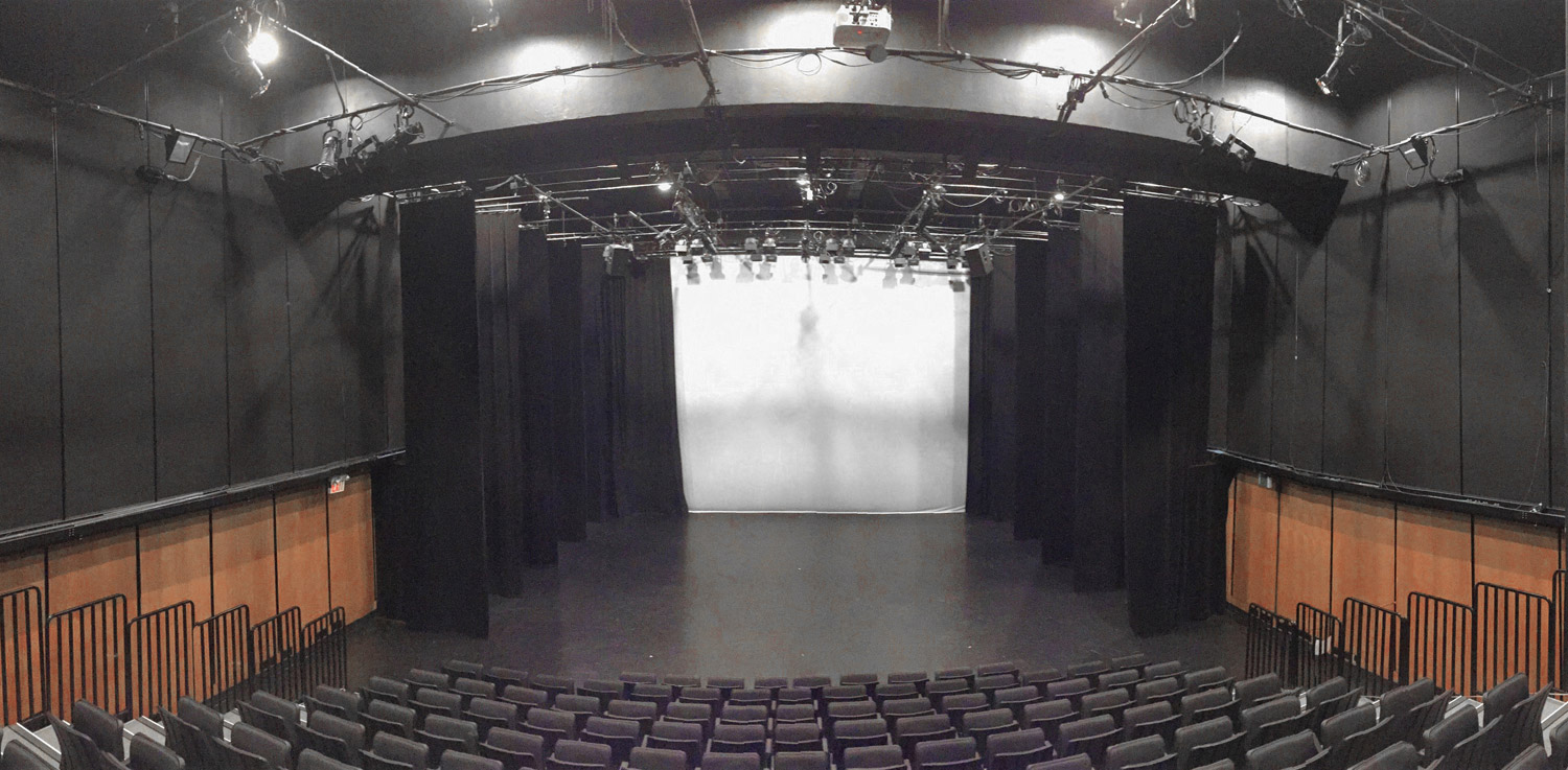 An empty black box theatre with folding seats and a white backdrop on the stage.