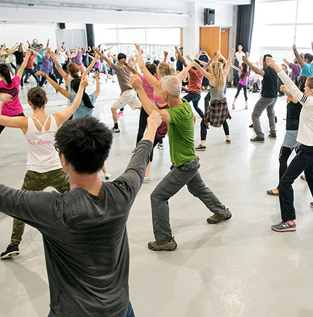 A group of people in an Open House dance class in a studio. They are wearing street clothes and have their arms extended above their heads in a V.