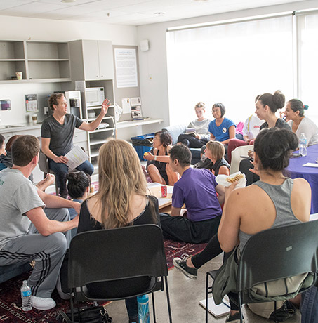 A blonde man in a black t shirt talks to a group of dancers sitting in an open space lounge area.
