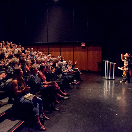 An audience watches a Greeting Ceremony in a black box theatre. A man in traditional Indigenous clothing and hat stands on the stage in front of a podium. He has one hand up in front of him and the other is holding a drum.