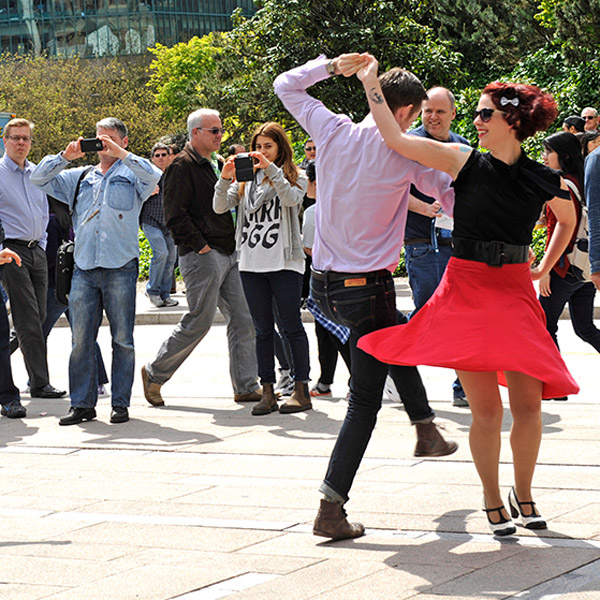 Two dance artists perform on sidewalk while people watch and take photos. The woman has a pink skirt that is twirling out and she moves.