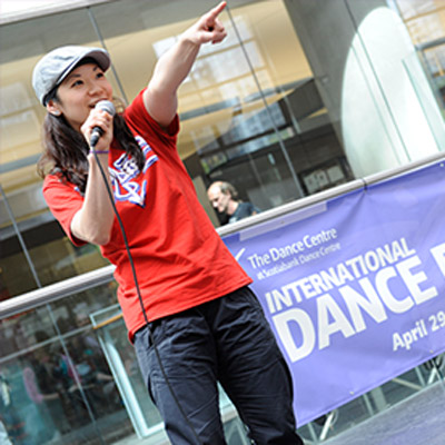 Woman pointing while speaking on microphone in the library lobby on International Dance Day. She has long brown hair and is wearing a red shirt and a baseball hat.