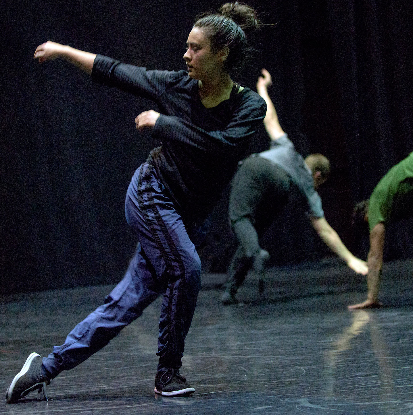 A dance artist poses mid curtsey with her arms outstretched. There are two other dance artists behind her on a black floor.