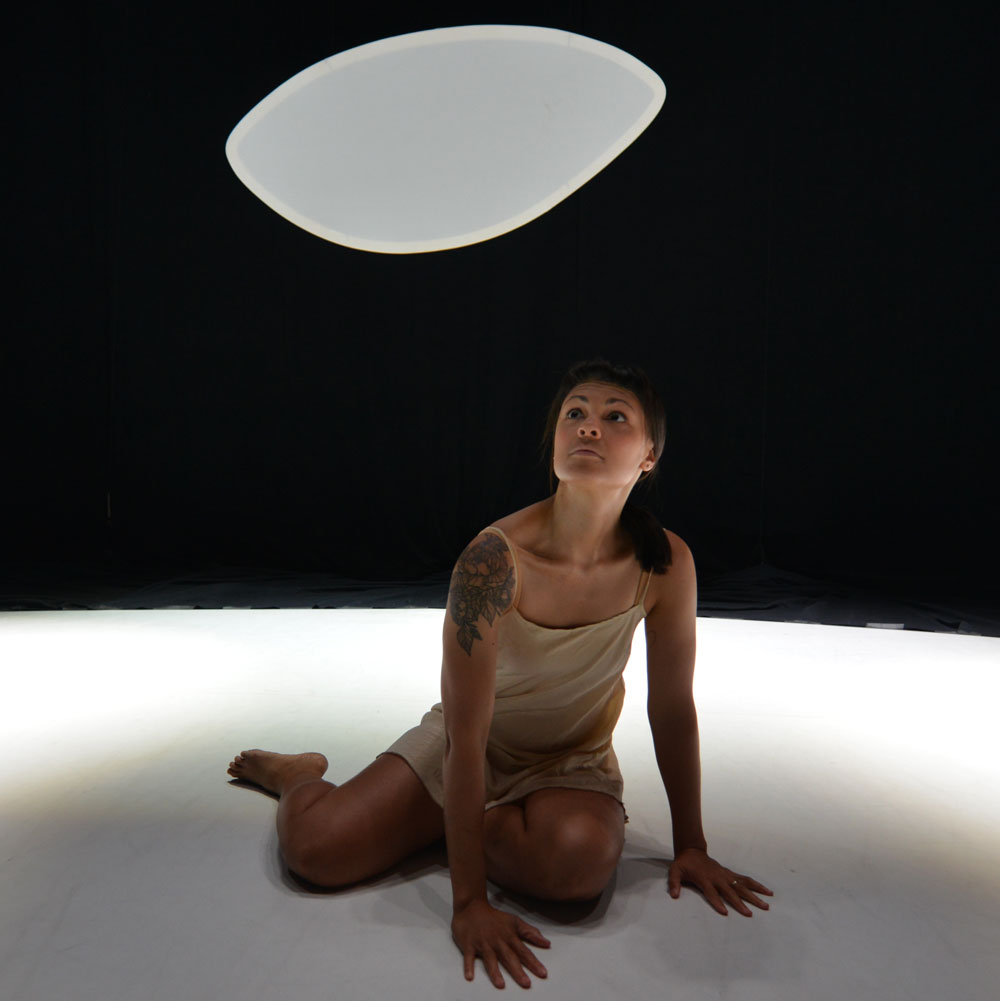 Amber Funk Barton sits on a white floor as she performs. There is a large white light hanging from the ceiling.