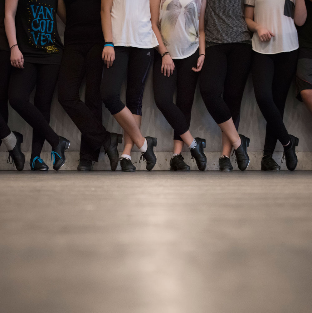 Many tap dancers stand against a wall in identical poses showing off their tap shoes