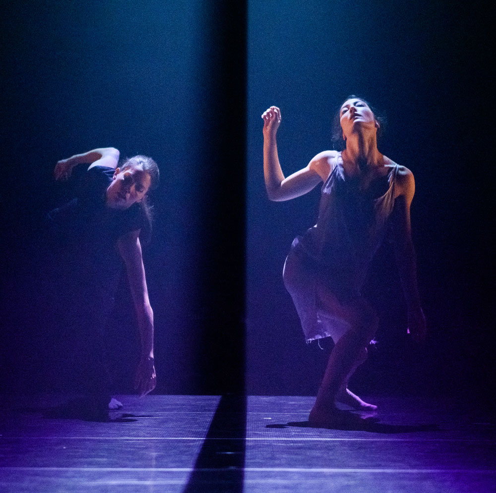 Two dance artists perform on stage. They are both partially bent over with spotlights on them.