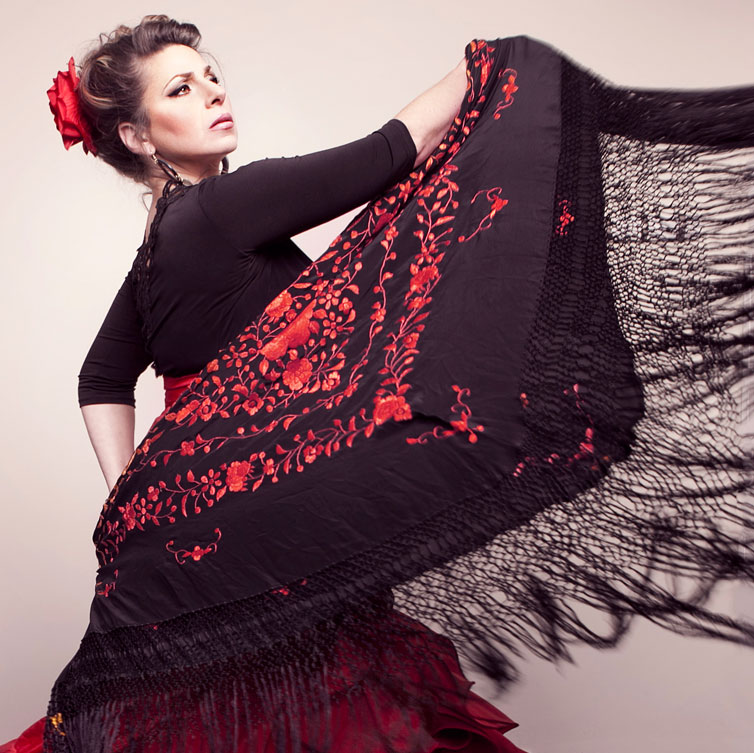 Flamenco artist Linda Hayes swings her arm out in front of her while holding her shawl. She is wearing a black top and a full red skirt. There is a red flower in her hair which is swept up. She also has long dangly earrings on.