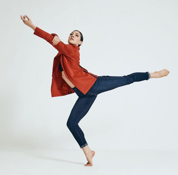Dance artist wearing blue jeans and a red shirt stands on one foot with the other leg extended behind her. Her left arm is also extended forwards.