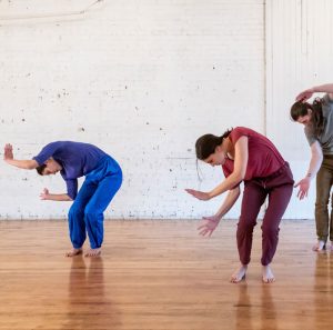 Three dance artists lean over toward their left with their arms outstretched while looking at the floor. They are wearing casual, bright, rehearsal clothing.