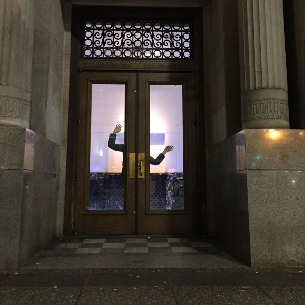 A dance artist poses behind the bank doors of Scotiabank Dance Centre with their arms outstretched to be seen out the door windows