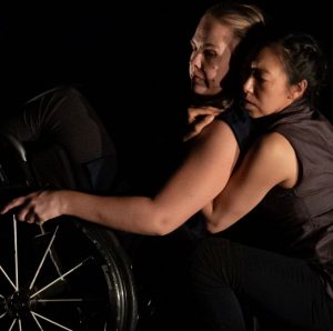 A dance artist in a wheelchair leans back and is held by another dance artist mid performance