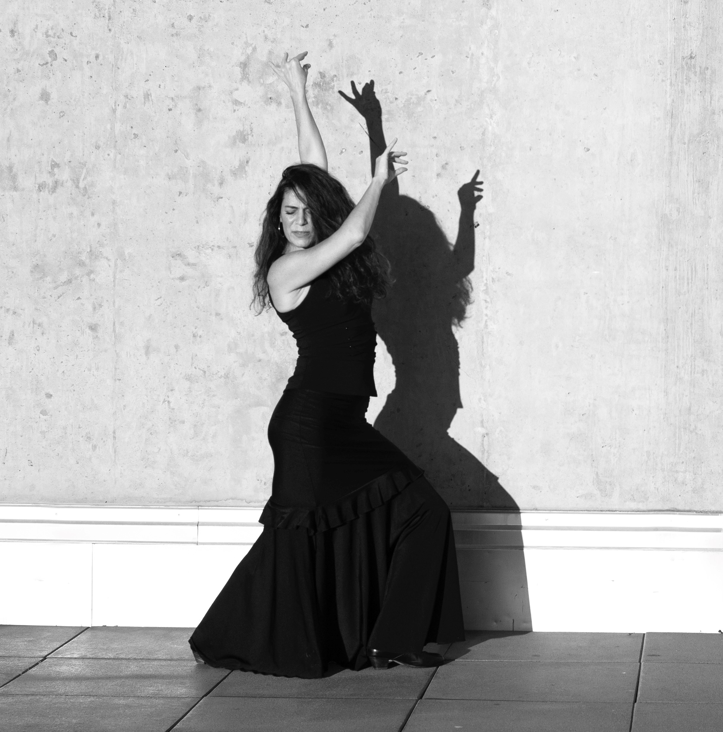 A flamenco dancer poses on a tile floor. She has a black dress on and her knees are bent with her arms extended above her head. The image is in black and white.