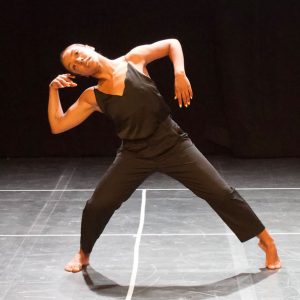 A female dance artist wearing a black jumpsuit poses with arms bent, leaning of her right leg