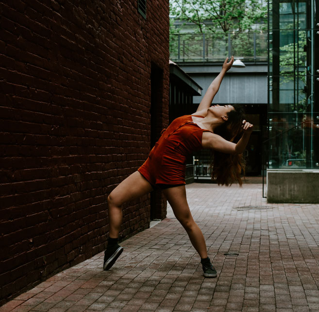 A dance artist wearing a red short jumpsuit poses in a cobblestone alley next to a brick wall. She is wearing black sneakers and is arched backwards with one knee slightly bent and her arms stretched over her head.