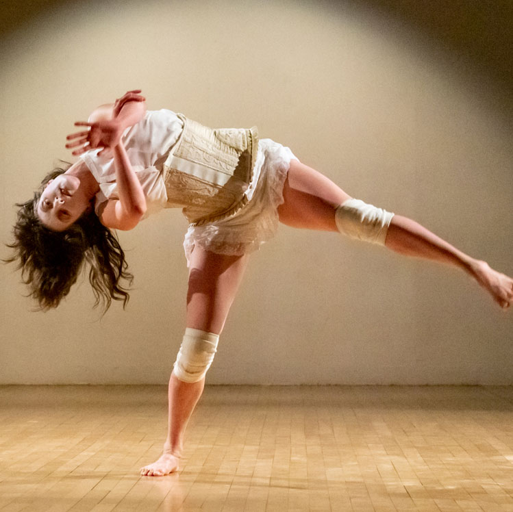 Dance artist Kate Franklin leans over on one leg, facing the camera with the other leg kicked out, and her arms pressed towards the camera