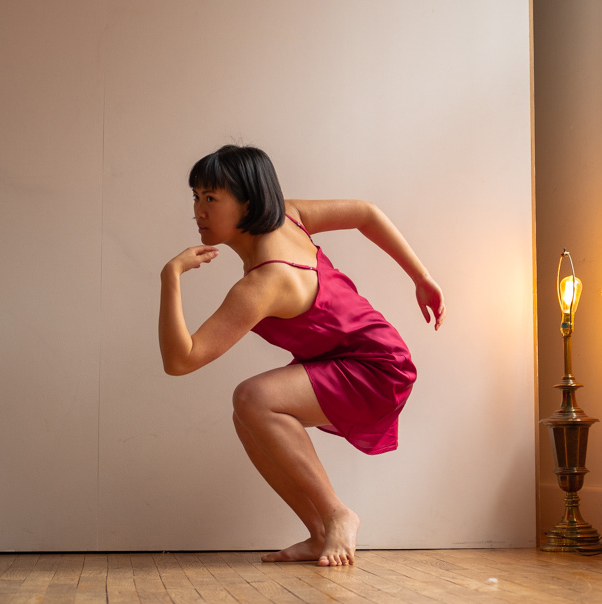 A dance artist wearing a pink dress crouches down with her left arm bent, touching under her chin. Her right arm is bent and extended behind her.