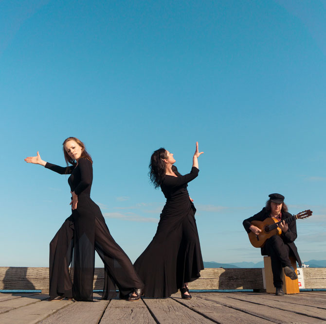 Two flamenco dancers pose on a pier. They are both wearing all black and are turned away from each other with one arm extended. There is a guitar player sitting on a box to the right of them in the photo wearing a black hat and suit.