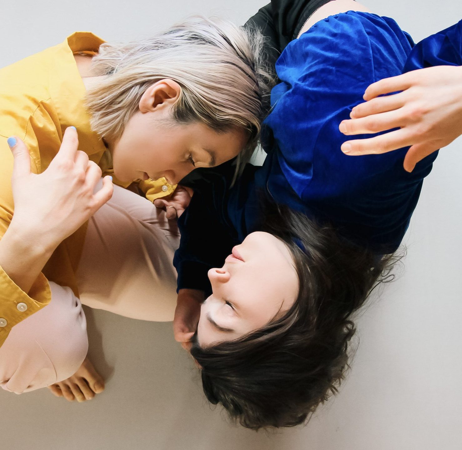 Two dance artists lean over with their heads towards each other. One is wearing a yellow shirt with blondeish hair and the other is wearing a blue shirt and has brown hair.