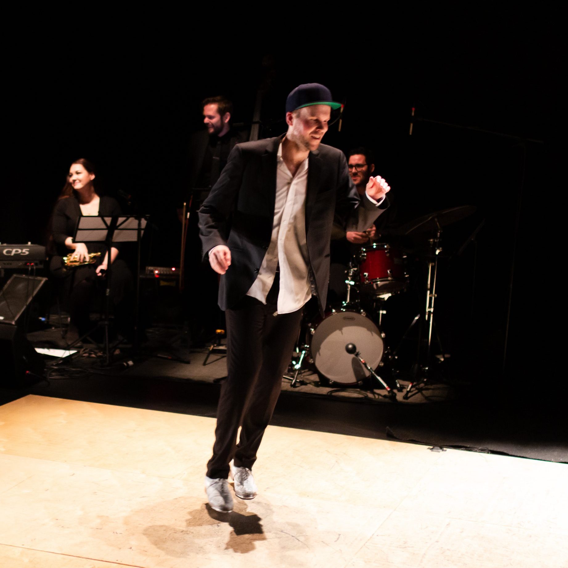 A tap dancer performs on a bright wood floor with a band behind him. He is wearing a ball cap and a black blazer and pants. The musicians behind him are laughing and playing in a casual manner.
