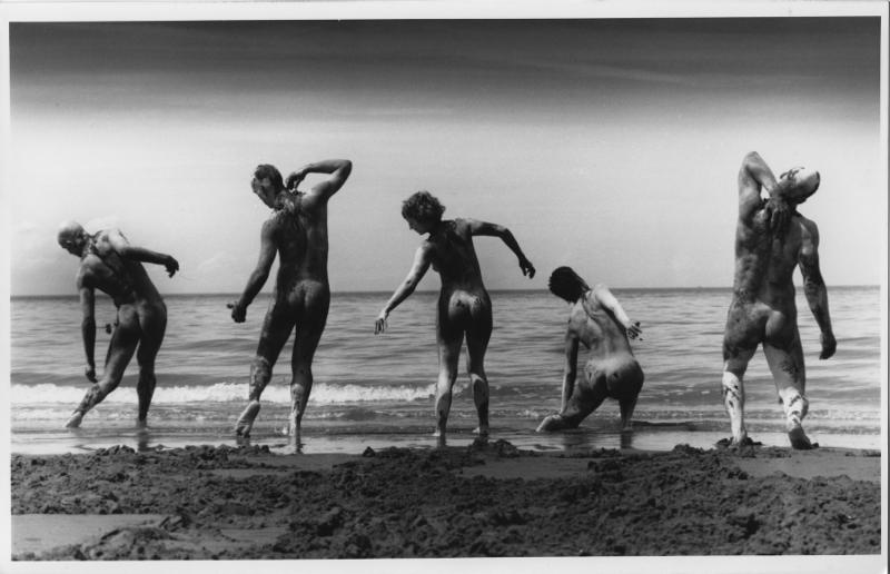 Five dancers pose mid performance on a beach. They are nude and facing the ocean, away from the camera. Their arms and legs are bent in various manners and one is kneeling in the waves.