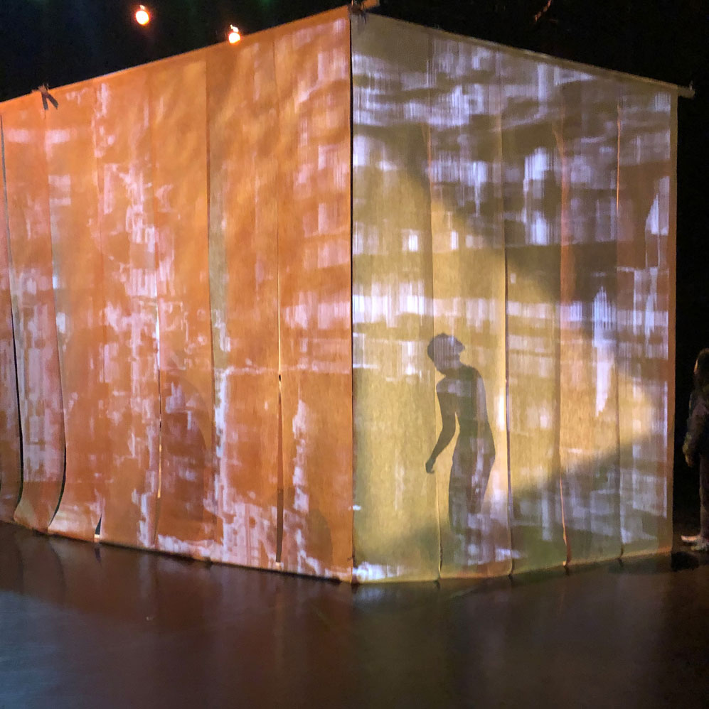 A large cube of paper stands in a black box theatre. It is lit from many sides, showing the silhouette of a dance artist standing on the inside of the box.
