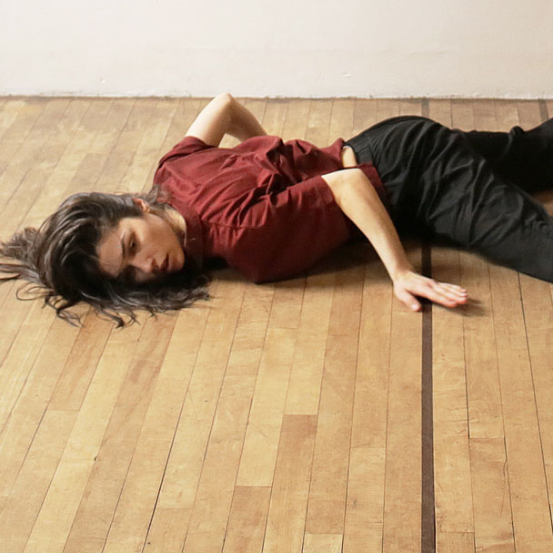 A dance artist lies on a wood floor. Her arms are bent up and she is wearing rehearsal clothes. Her brown hair is splayed out around her head.