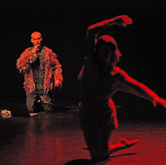 Two people on a dark stage lit by a red light. In the background a man kneels with a microphone held to his mouth and a jacket made of shaggy fabric. In the foreground, out of focus, a woman also kneels with her arms outstretched.