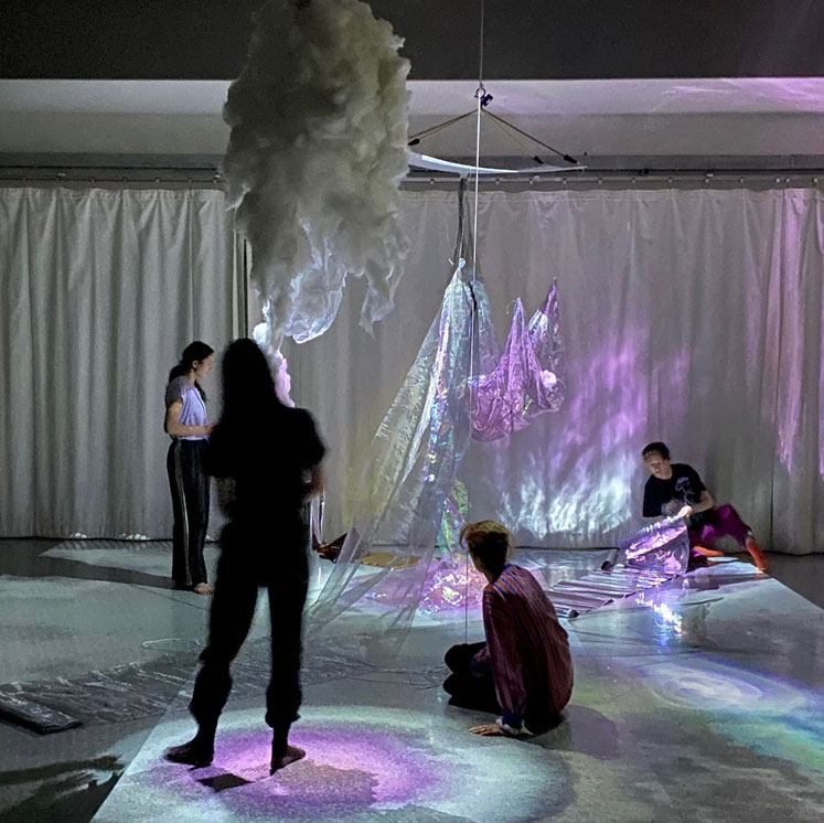 Four dance artists sit or stand looking at an installation in a dance studio. There is fluffy cloud like material hanging from the ceiling and shiny purple fabric draped up a rope as well. The floor is lit with purple swirly lights.