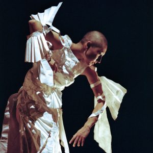 A dance artist leans over on a black stage with her arms outstretched. She is wearing a white dress and fan like wings on her elbows.