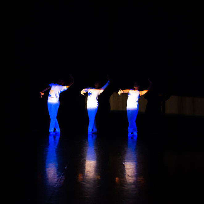 Three dance artists perform on a dark stage. They are wearing white clothing and there is a bright spotlight shining on their torsos, obscuring their faces. Their right arms are reaching out in the front of them in the spotlight, their left arms are reaching up above them in the dark.