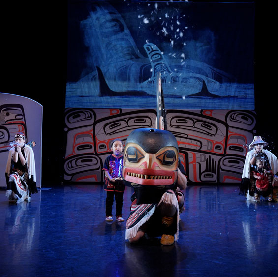 Three Indigenous dance artist pose on stage. Two in the background are wearing traditional clothing and head pieces, the one in the front holds a large whale head in front of their body. There is a small child next to them looking out towards the audience. Behind the artists are backdrops painted with tradition art and a screen projecting images of Orca swimming overlapped with traditional drawings of Orca.