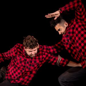 Two artists on a black stage. They are both wearing red and black flannel shirts. One, with curly brown hair, is crouched down with his left arm pushing into the stomach of the other artist who is crouching above him with one arm bent up above his arm.
