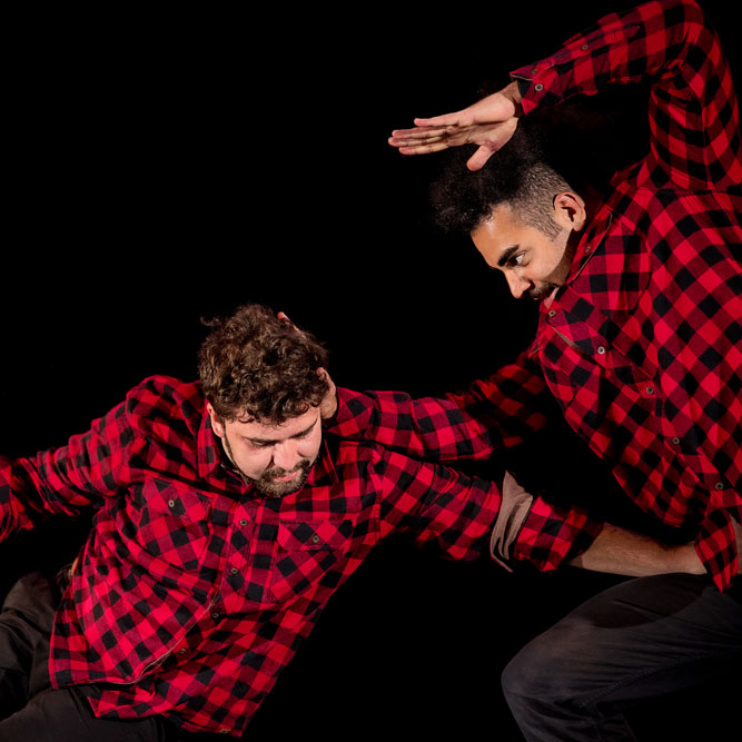 Two artists on a black stage. They are both wearing red and black flannel shirts. One, with curly brown hair, is crouched down with his left arm pushing into the stomach of the other artist who is crouching above him with one arm bent up above his arm.