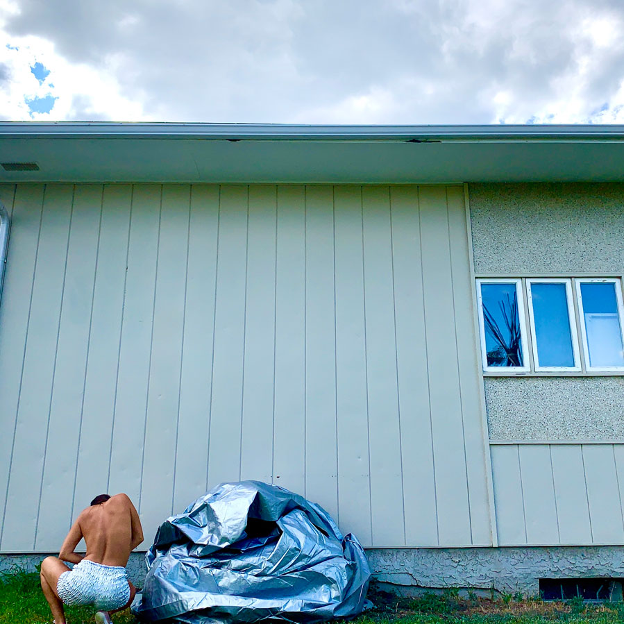 A dance artist crouches down away from the camera, facing a clapboard house. They are wearing short frilly white shorts and their top is bare. Next to them is a silver fabric arranged in a circular pile. They are crouching on grass and there is clouds in the sky.