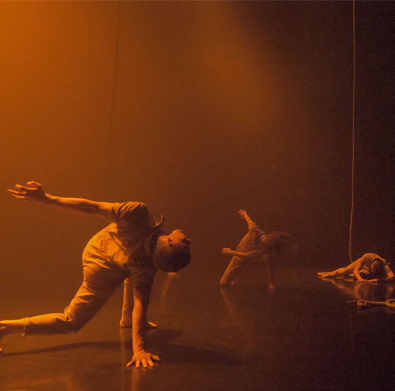 Four dance artists crouch in various positions on a smoky orange stage. They are wearing beige clothing and there are three ropes hanging from the ceiling