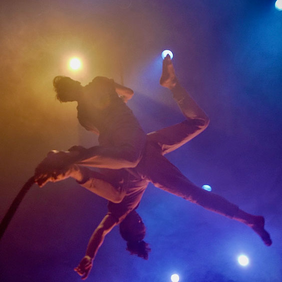 Two dance artists hang from a rope. The air around them is hazy. One artist hangs upwards with their back arched and knees bent backwards. The other has their back pressed up against them and is lying perpendicular to the first artist. They have one leg bent, their arms outstretched and their head is looking down towards the floor.