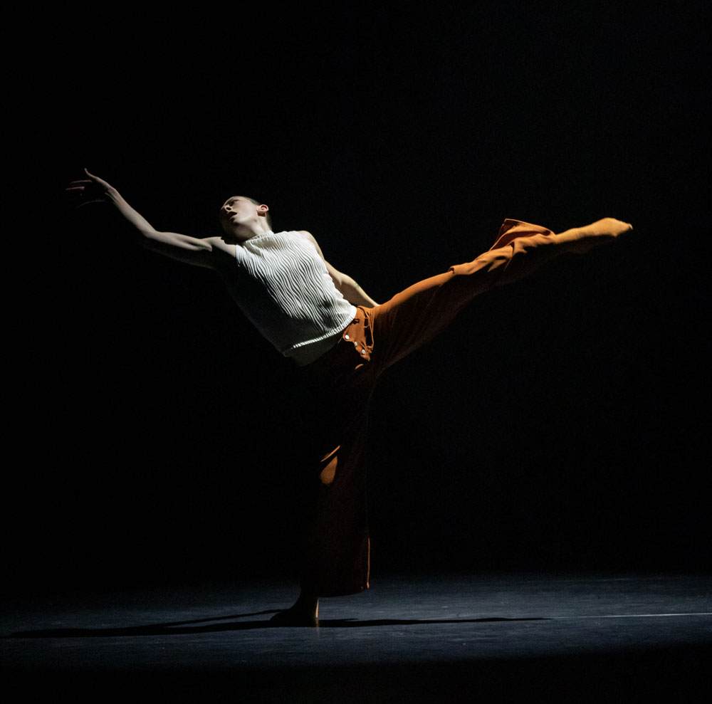 A dance artist on a black ground. Their left leg is outstretched as is their right arm and their head is tilted back. They are wearing a sleeveless white sweater and orange pants and socks.