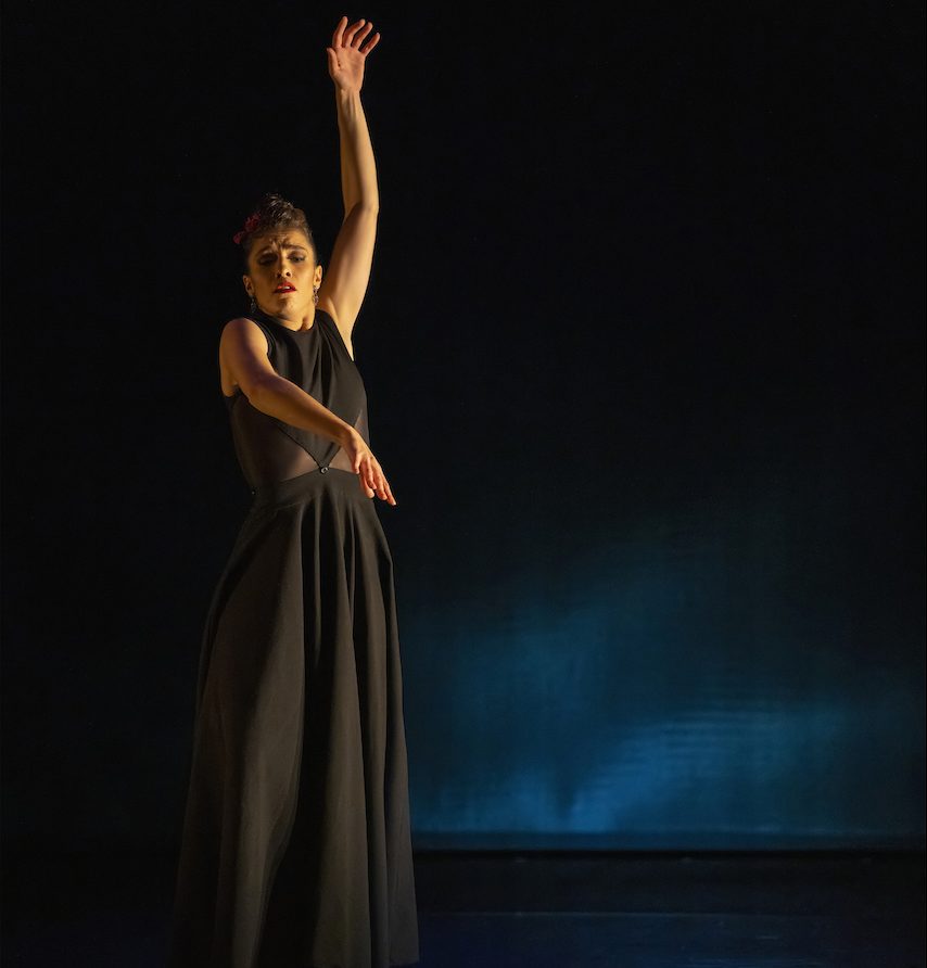 A dance artist stands on a black stage. There is a blue light reflecting from the floor on to the back wall. The artist is wearing a long black sleeveless dress and has one arm out in front of them and the other up above their head. They have dark hair, pulled back, and bright red lipstick.