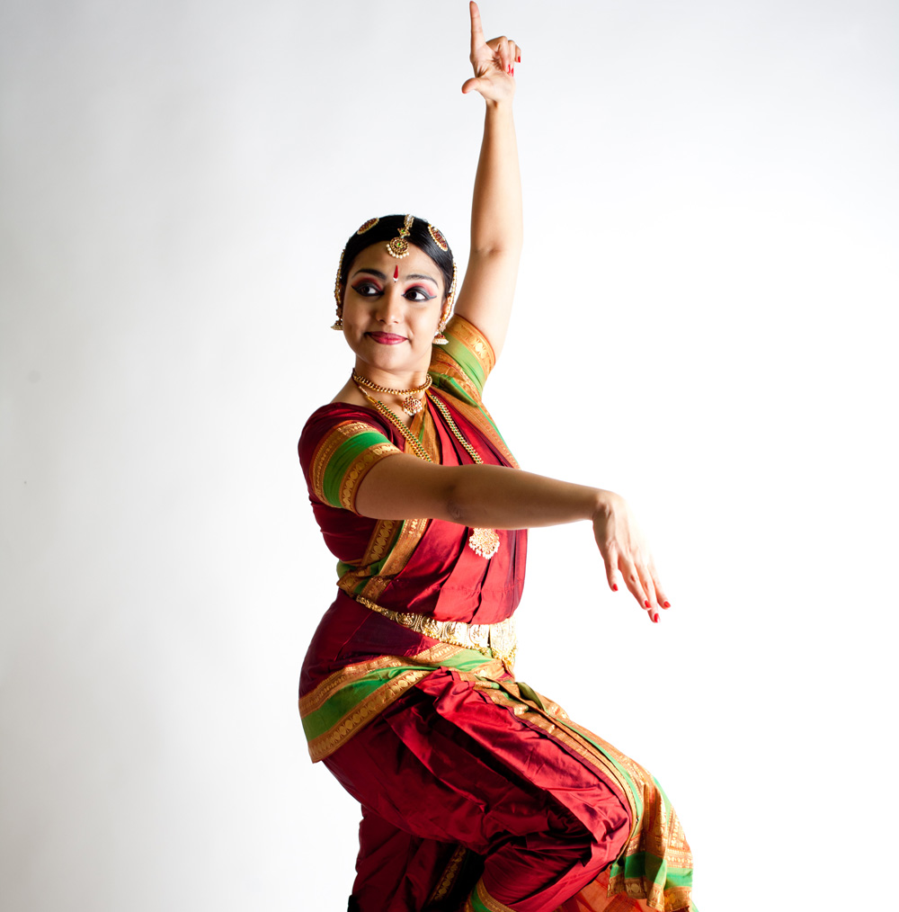 A dance artist poses in a traditional bharatanatyam outfit. Their left arm is pointed upwards and their right knee is bent up to rest of their leg.