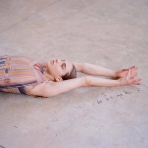 A dance artist lies on concrete with their arms stretched up above their head.