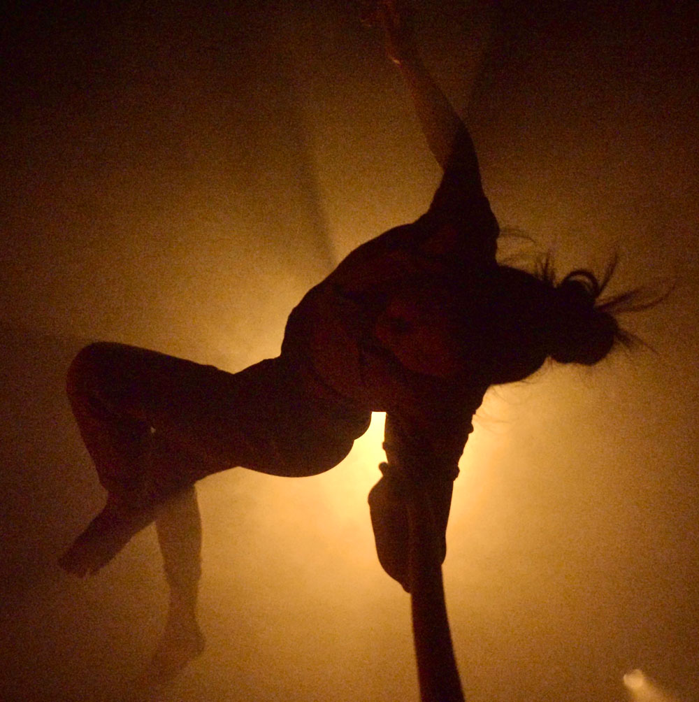 A dance artist hangs in the air with their head pointed down, towards the camera. Their dark hair is in a bun and they have their arms extended to the sides. There is a bright light just behind them giving the image an orange tint.