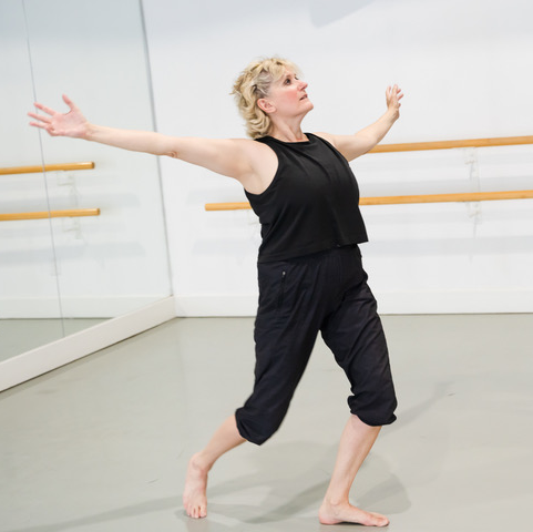 A dance artist poses in a bright studio. They are wearing all black - capris and tank top. They have short blond hair and have their arms extended outwards.