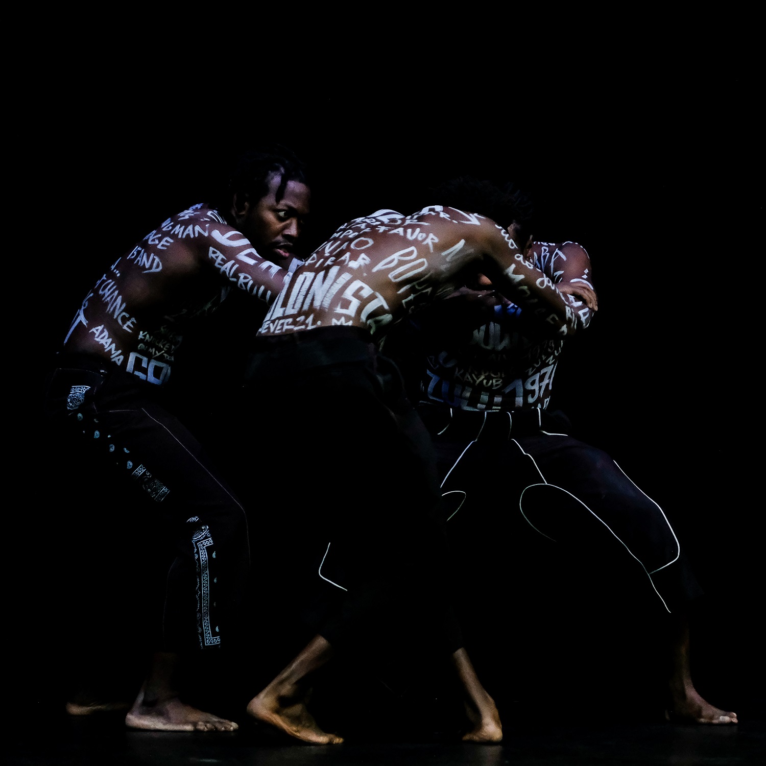 Three dance artists bend over in towards each other on a black stage. They are topless with words written in white paint all over their torsos and arms. They have short black pants and bare feet.