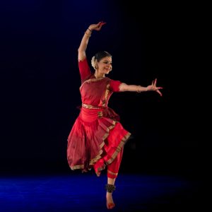 A dance artist poses mid performance on a dark stage. They are wearing a full bharatanatyam costume and head dress. Their toes and fingers are painted red and one leg is bent up behind them. Their arms are outreached over their head and to the side.