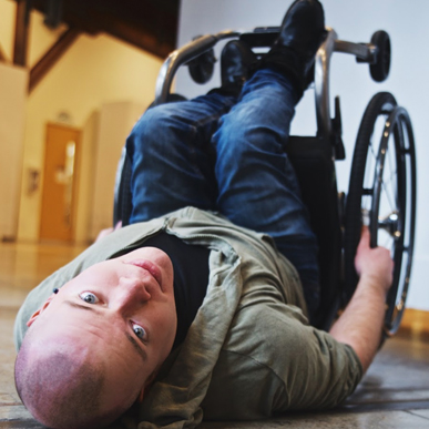 A seated dance artist lies on a floor looking at the camera. His legs and chair are being held up perpendicular.