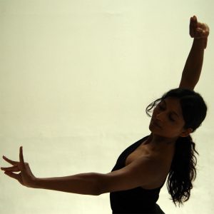 A dance artist poses with one arm out to the side and the other above their head. They are wearing a one arm black shirt and have a long black curly ponytail. Their head is tilted to the left and their eyes are closed. Their fingers on both hands have their index finger and thumb pressed together.